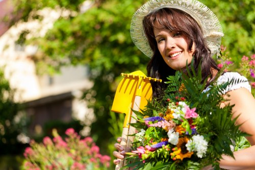 A woman in a white hat with a wide brim is standing in a well-maintained garden, holding a vibrant bouquet of mixed flowers and greenery. The garden features a lush green lawn in the foreground, bordered by flower beds with colorful blooms, and a background of tall, leafy trees providing shade. The scene suggests a sunny day with natural lighting highlighting the vivid colors of the flowers and the greenery. The garden appears tidy and landscaped, suitable for outdoor gardening and maintenance services offered by Garden Maintenance Ealing, reflecting a peaceful and inviting outdoor space in Londonâ€™s Ealing area.